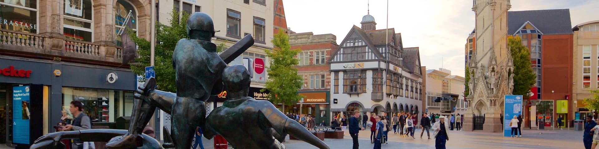 A statue of two figures in a square with historic buildings and a clock tower in the background.