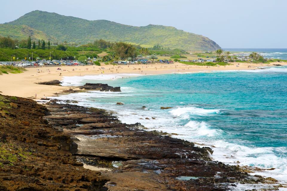 A coastal beach with rocky cliffs, clear turquoise waters, and a mountainous backdrop.