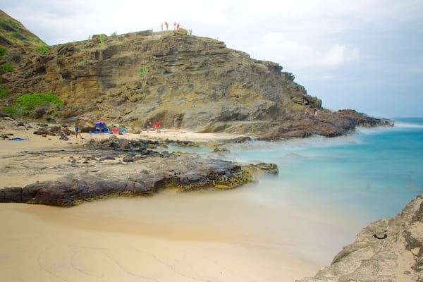 Une plage rocheuse bordée d’une eau turquoise et limpide, une étendue de sable et une falaise sur laquelle se tiennent des personnes.