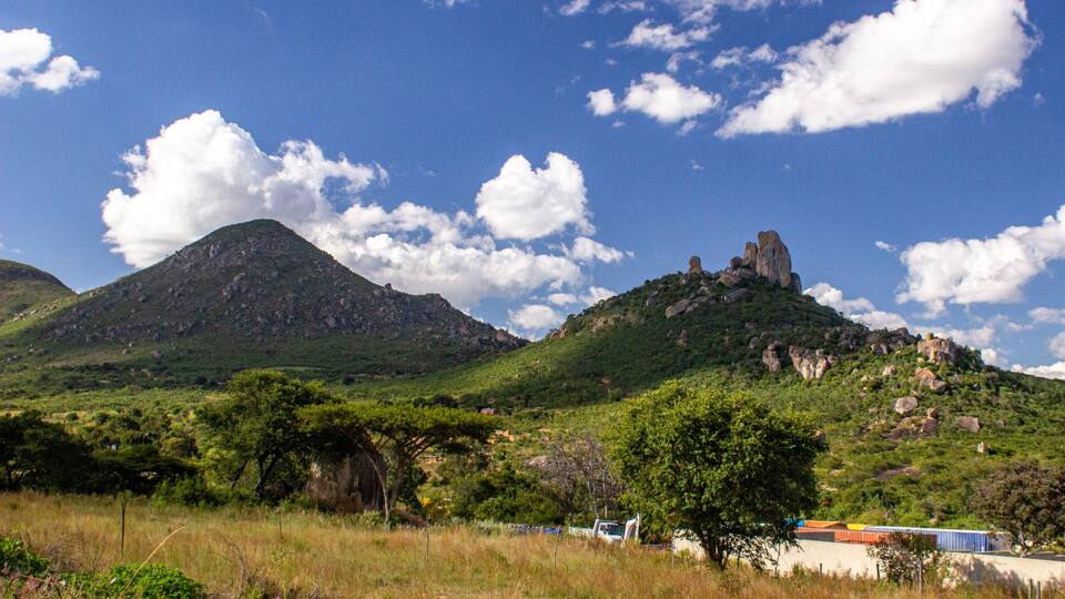 A mountainous landscape with a prominent rock formation on a hill, surrounded by greenery and a clear blue sky with scattered clouds.