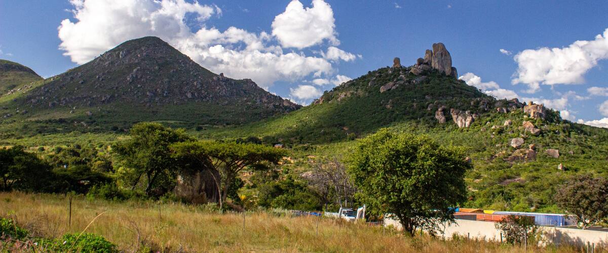 A mountainous landscape with a prominent rock formation on a hill, surrounded by greenery and a clear blue sky with scattered clouds.