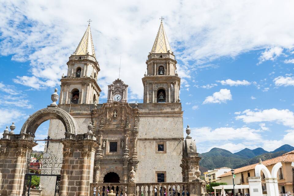 Una catedral histórica con dos torres, un arco ornamentado y un paisaje montañoso de fondo.