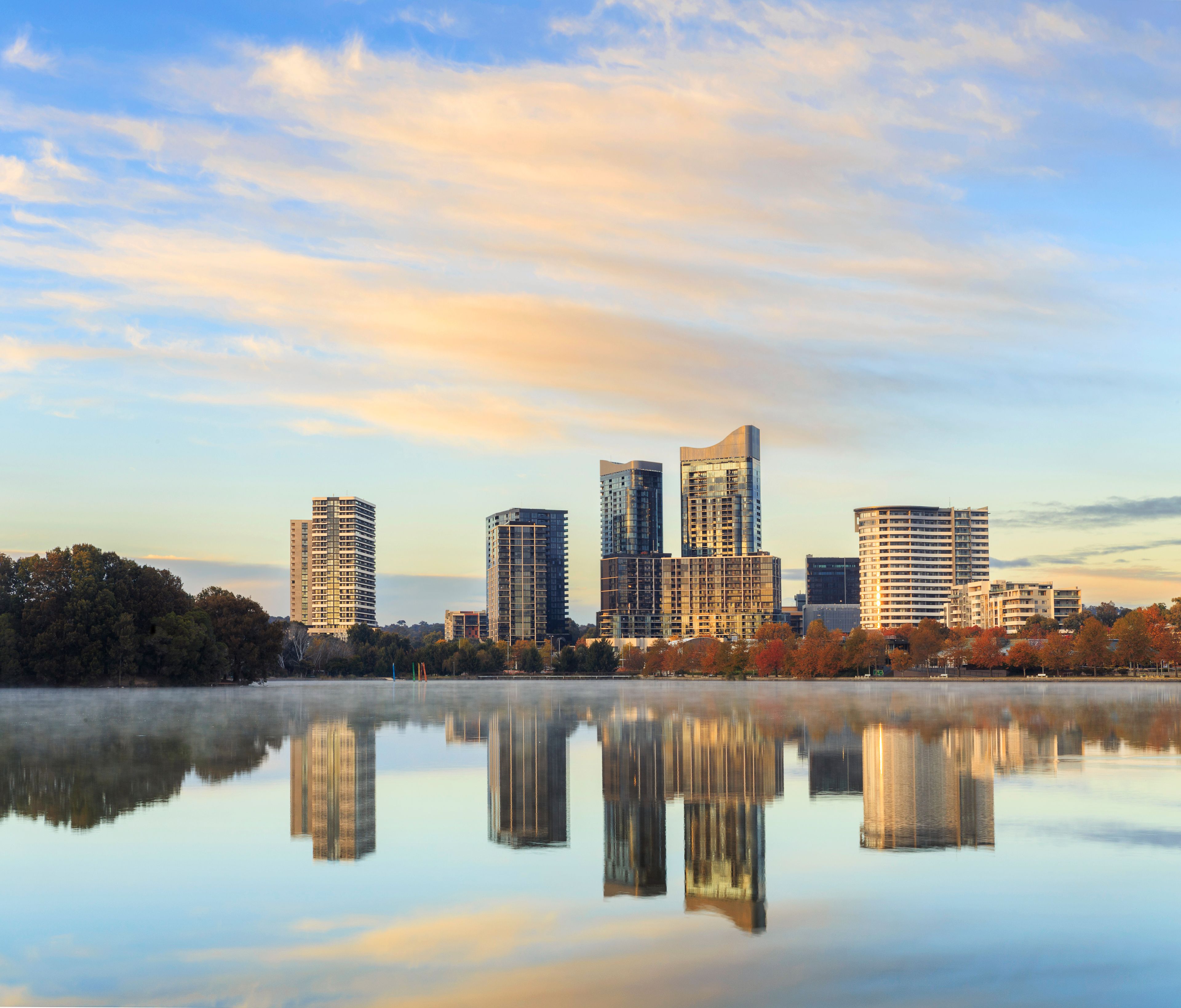 A city skyline with modern buildings reflected in a calm body of water, surrounded by trees with autumn foliage.