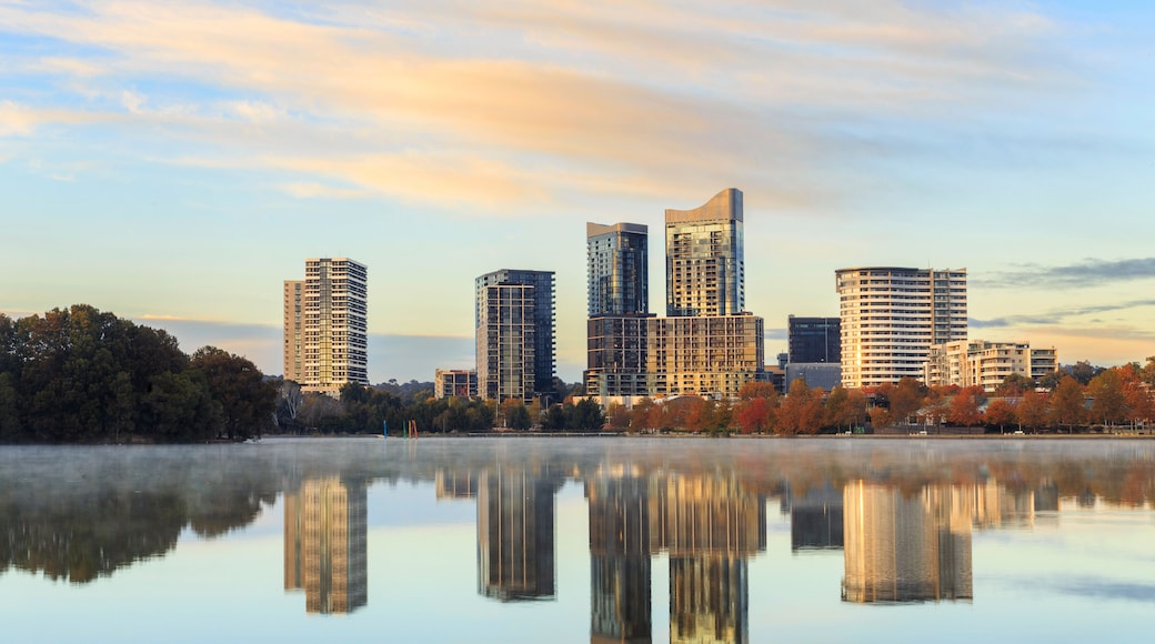 A city skyline with modern buildings reflected in a calm body of water, surrounded by trees with autumn foliage.