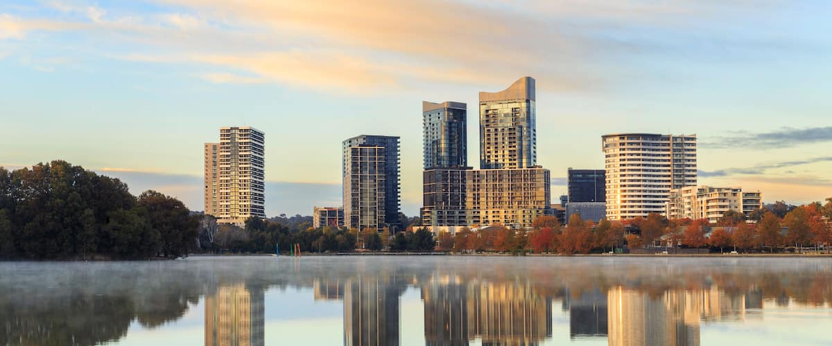 A city skyline with modern buildings reflected in a calm body of water, surrounded by trees with autumn foliage.