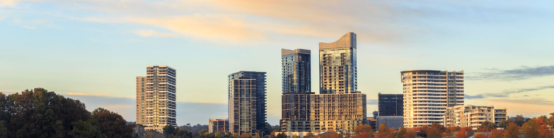 Silhueta urbana com edifícios modernos refletidos em águas calmas, cercada por árvores com folhagem de outono.