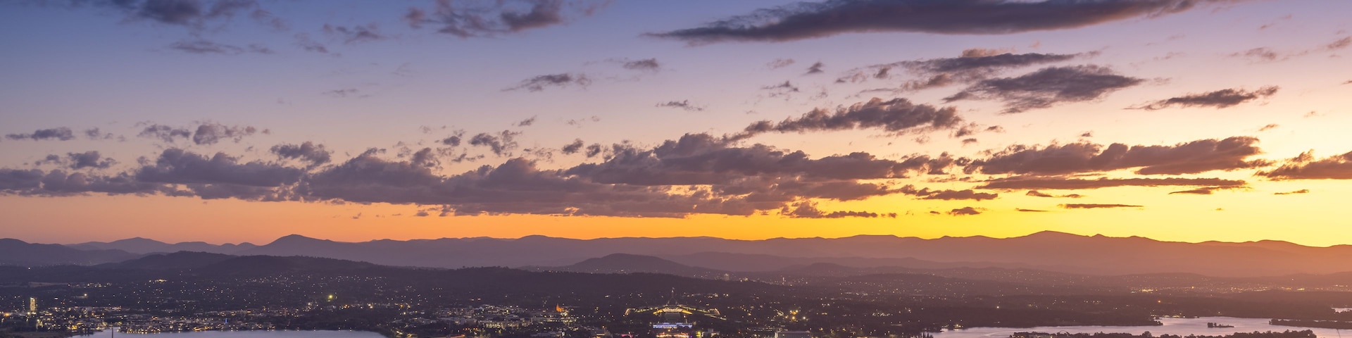 Paisagem urbana ao entardecer, com um rio, montanhas ao fundo e uma ponte.