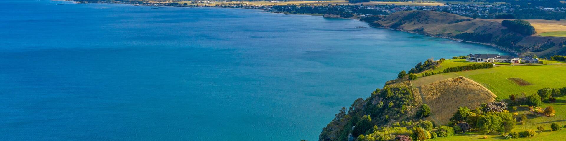 A coastal landscape with a large body of water, green hills, and a distant mountain range under a partly cloudy sky.