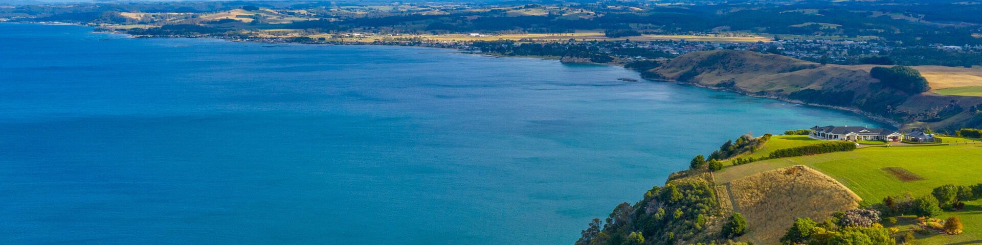 A coastal landscape with a large body of water, green hills, and a distant mountain range under a partly cloudy sky.