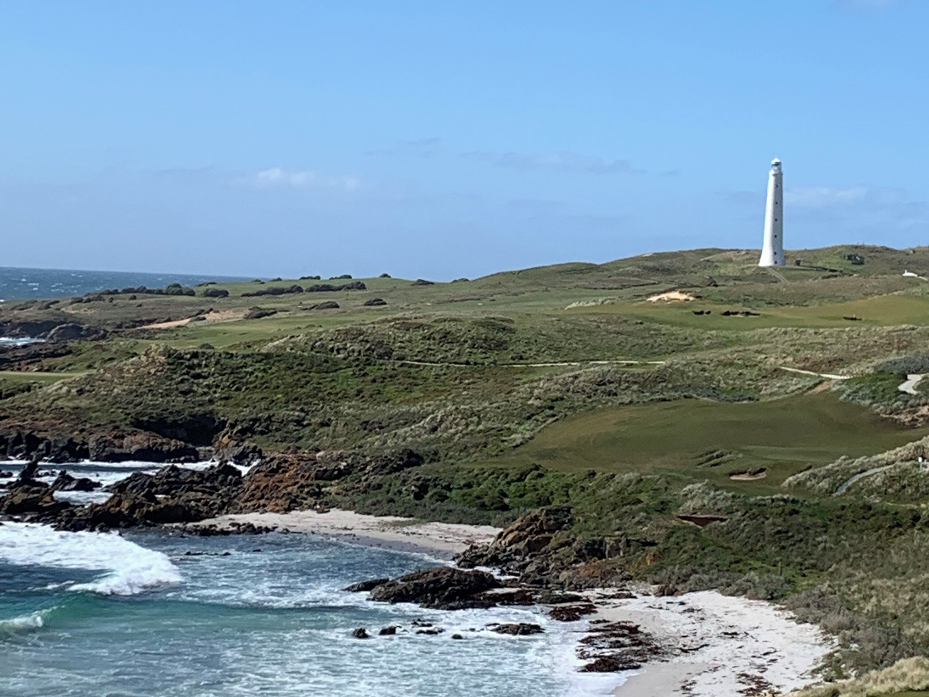 A coastal landscape with a lighthouse on a grassy hill, rocky shoreline, and waves crashing onto the beach.