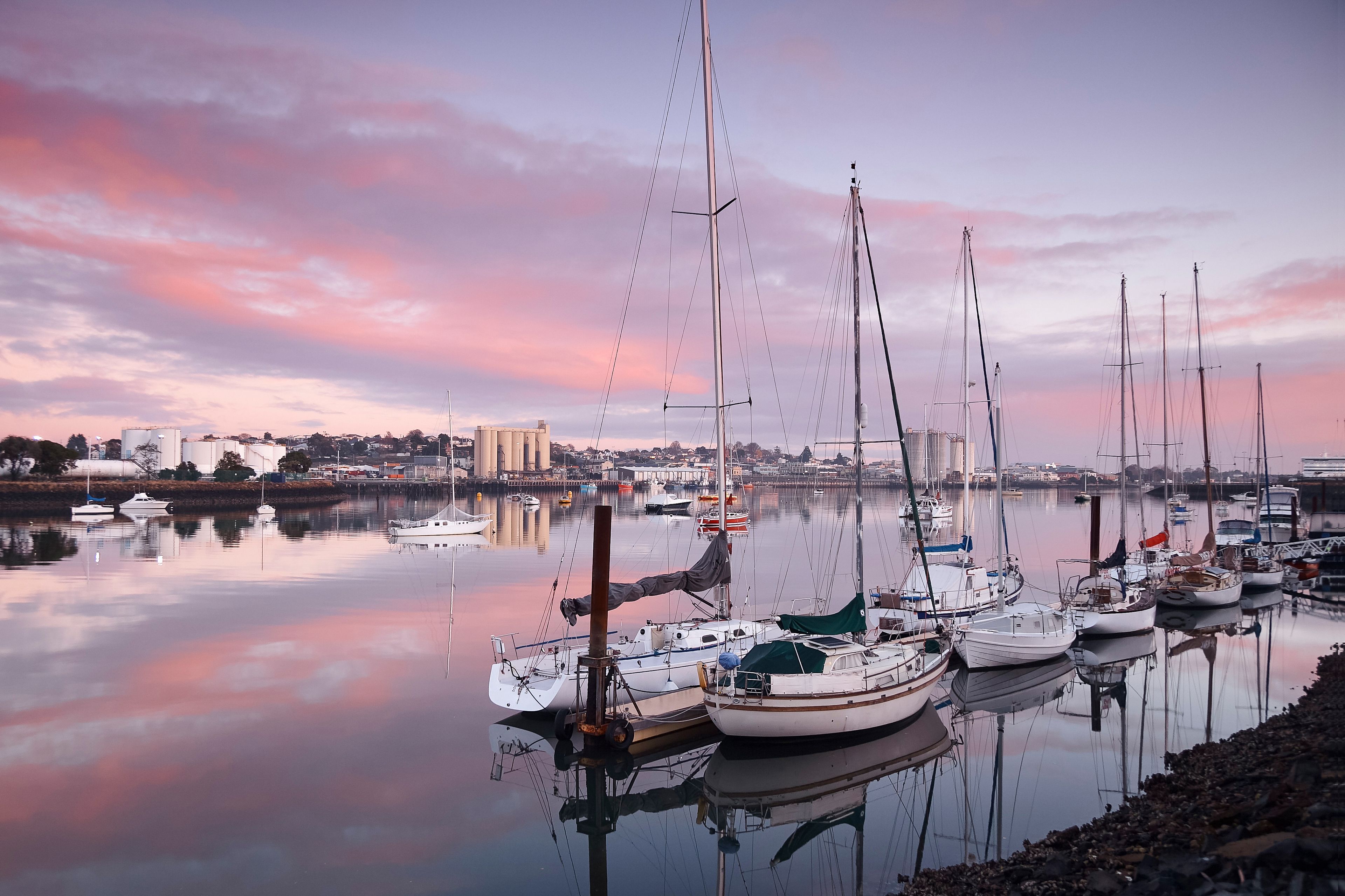 Un puerto deportivo con varios veleros amarrados, una superficie acuática tranquila que refleja el cielo, y un paisaje urbano al fondo.