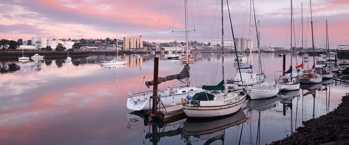Un port de plaisance où sont amarrés de nombreux voiliers, une étendue d’eau calme qui reflète le ciel et, au loin, une vue sur la ville.