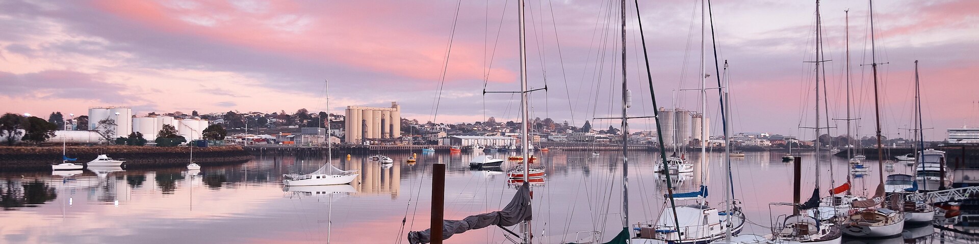 Un puerto deportivo con varios veleros amarrados, una superficie acuática tranquila que refleja el cielo, y un paisaje urbano al fondo.