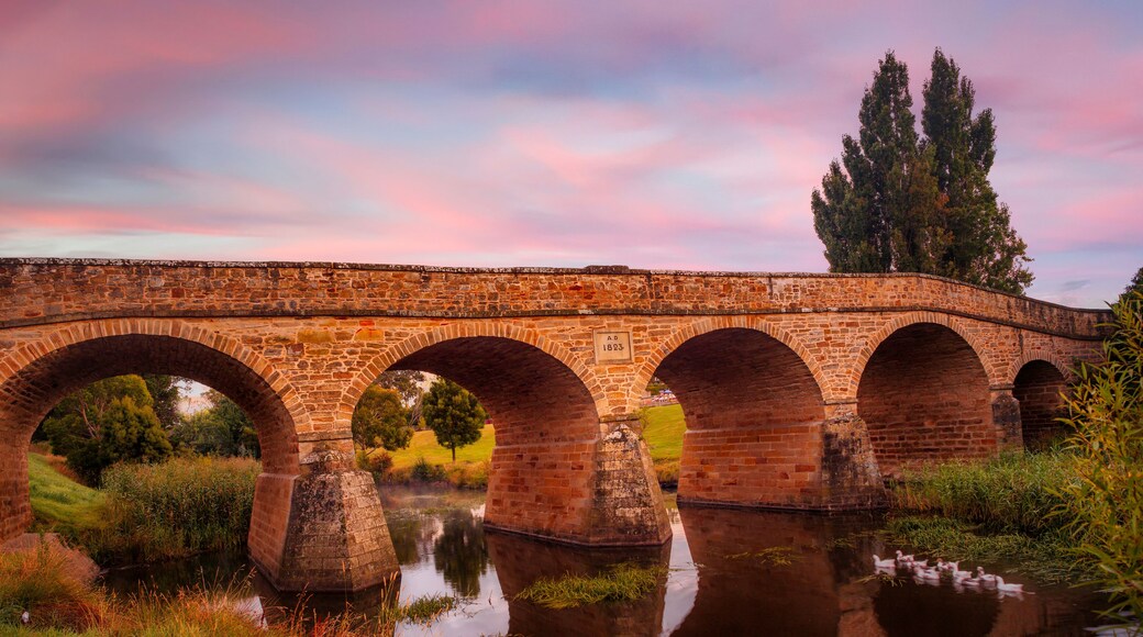 A stone bridge with multiple arches over a calm river, surrounded by greenery and trees.