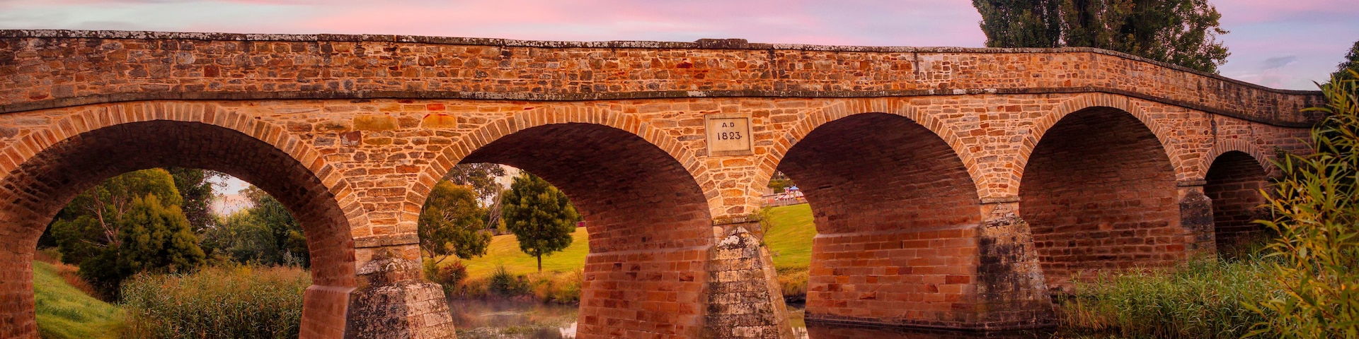 A stone bridge with multiple arches over a calm river, surrounded by greenery and trees.