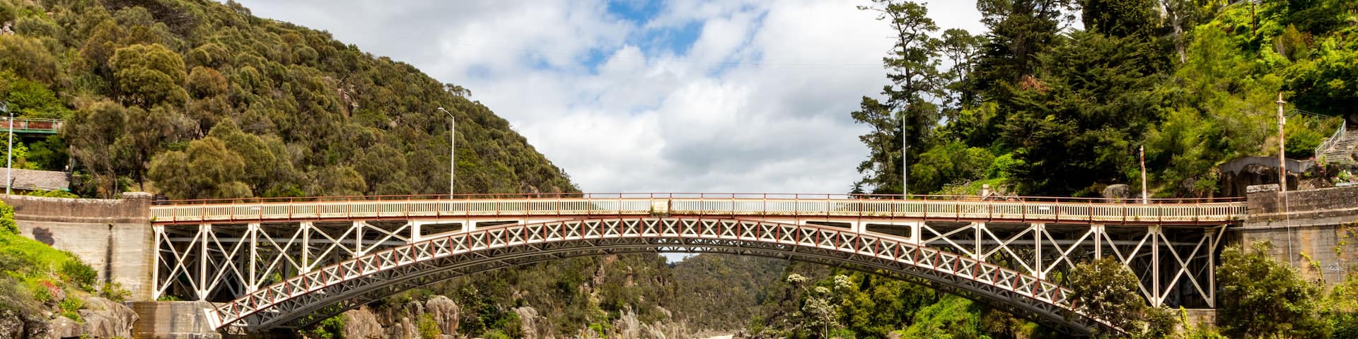 A steel bridge over a river with surrounding greenery and hills.