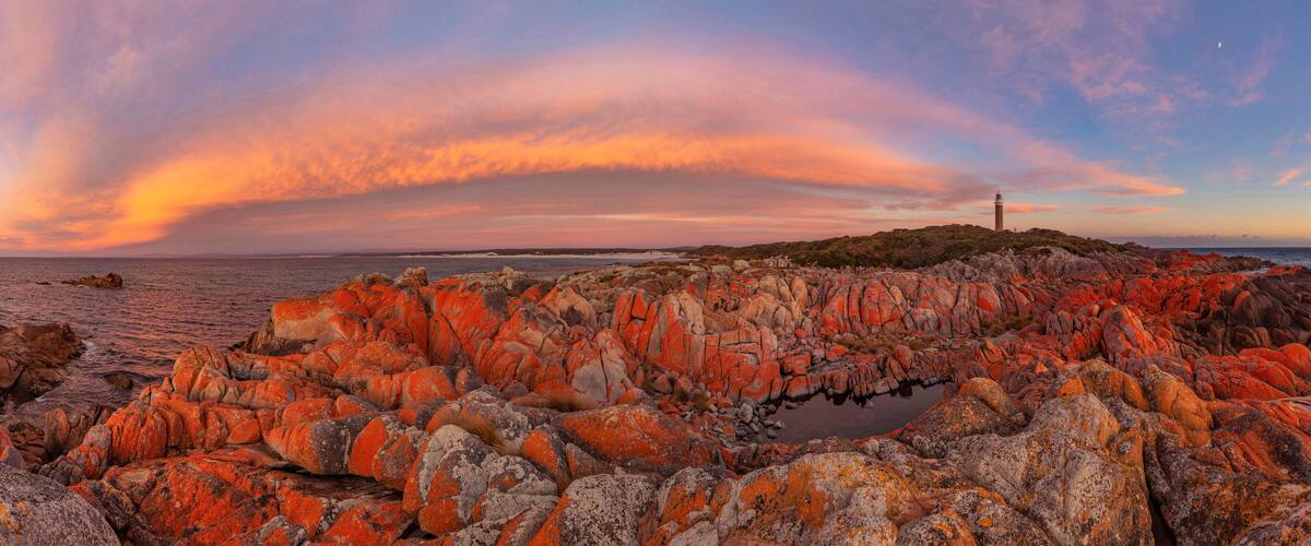 A coastal landscape with striking red rocks and a lighthouse in the distance.