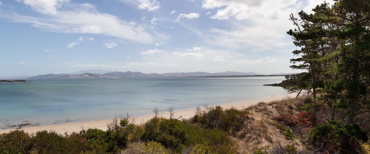 A coastal landscape with a sandy beach, calm sea, and distant mountains under a partly cloudy sky.