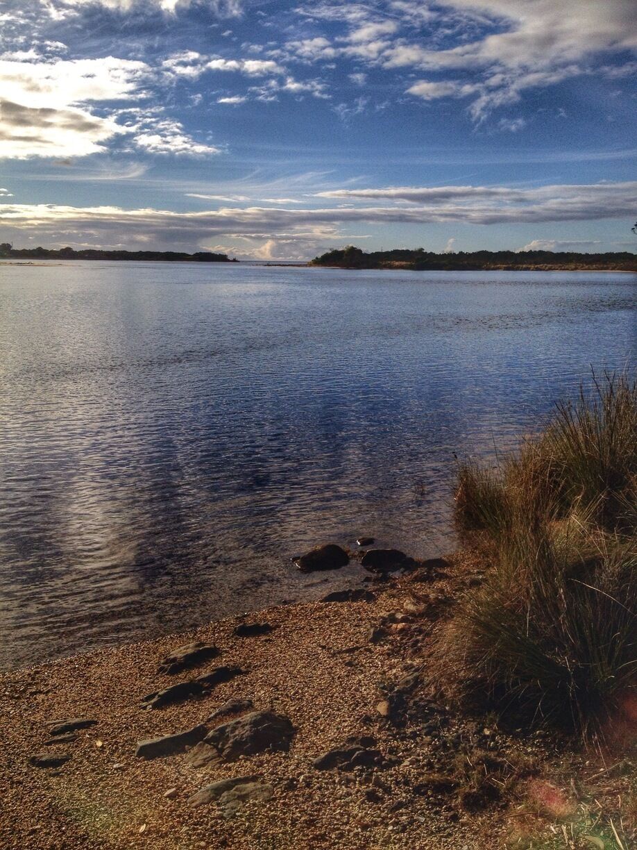 A calm lake with pebbles and grass along the shore, a distant shoreline, and a partly cloudy sky.