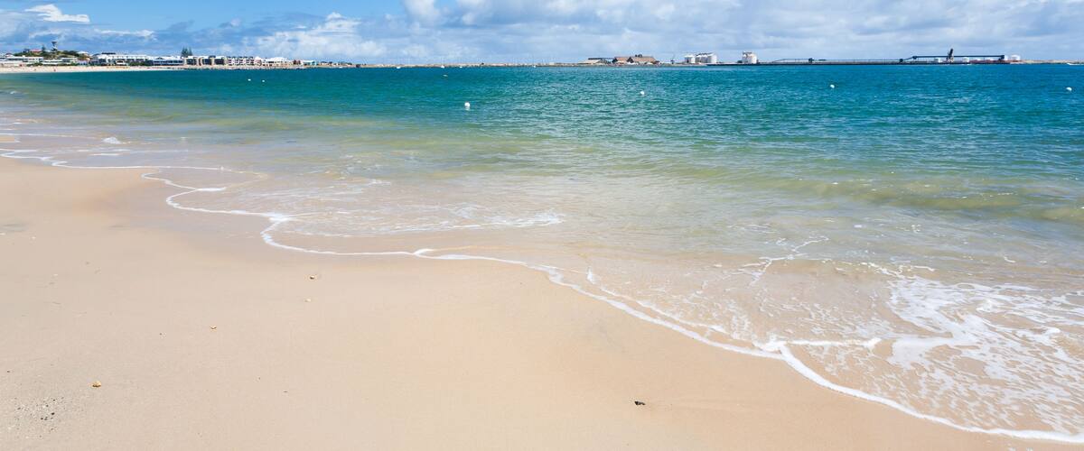 A beach with clear turquoise water, gentle waves, and a distant bridge.