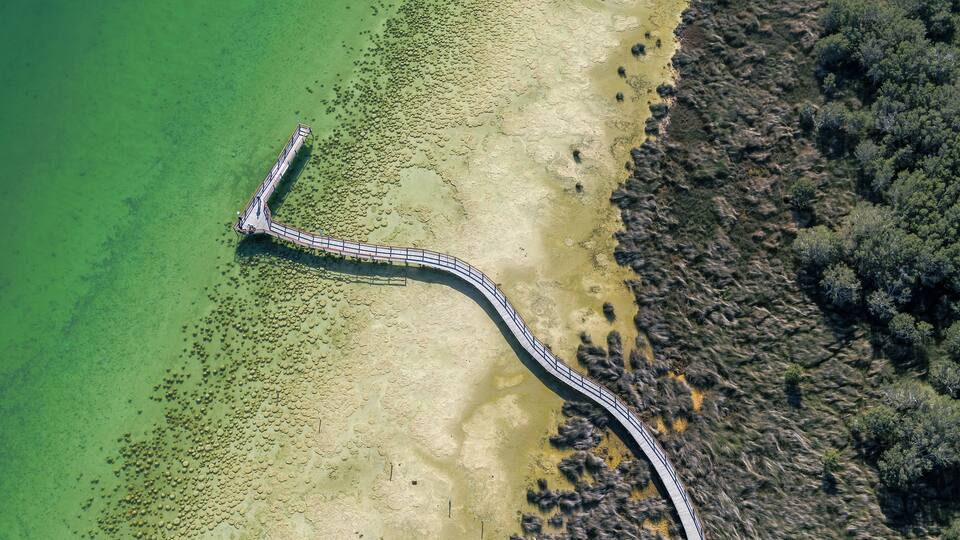 An aerial view of a curved bridge over a shallow, rocky water body with clear green water, leading to a forested area.