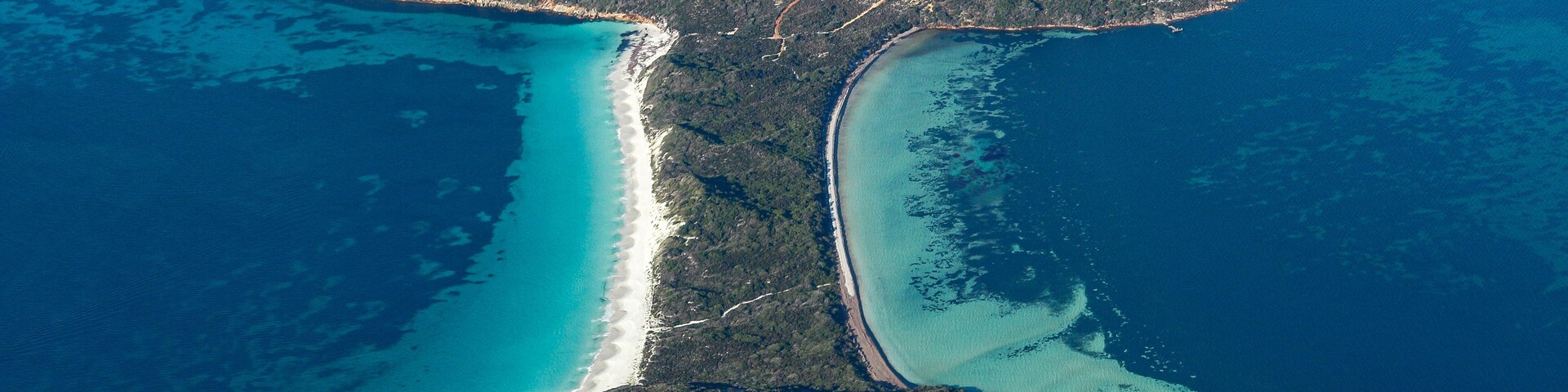 Vista aérea de uma área costeira com um istmo estreito ligando duas massas de terra maiores, cercada por águas turquesa e vegetação densa.