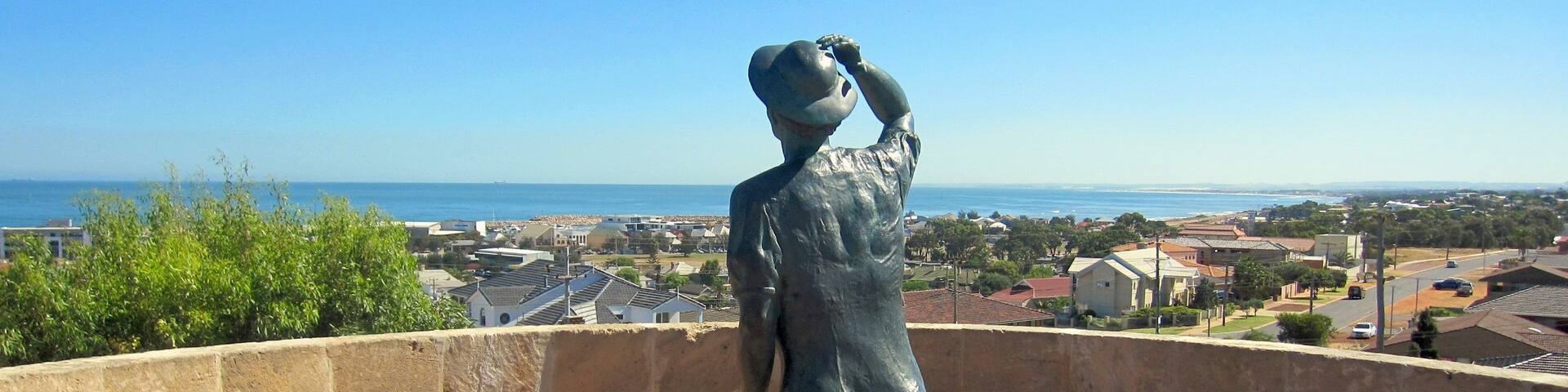 A statue of a woman in a dress standing on a brick platform overlooking a coastal town.