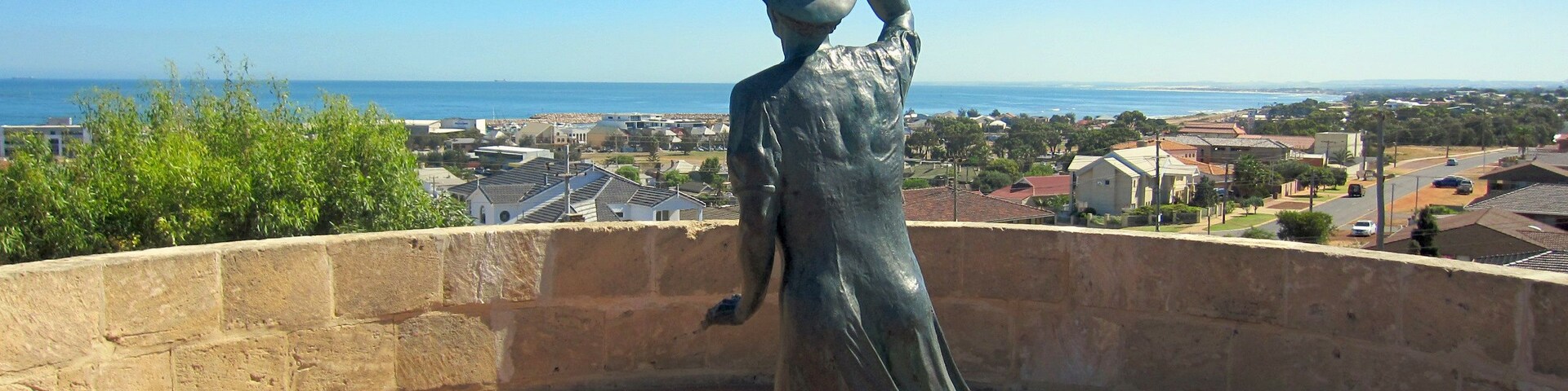 Una estatua de una mujer con vestido, sobre un pedestal de ladrillo, con vista a una ciudad costera.