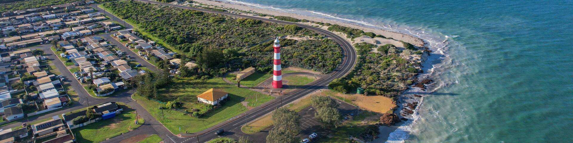 An aerial view of a coastal area with a lighthouse, a curved road, and a residential area with houses and greenery.