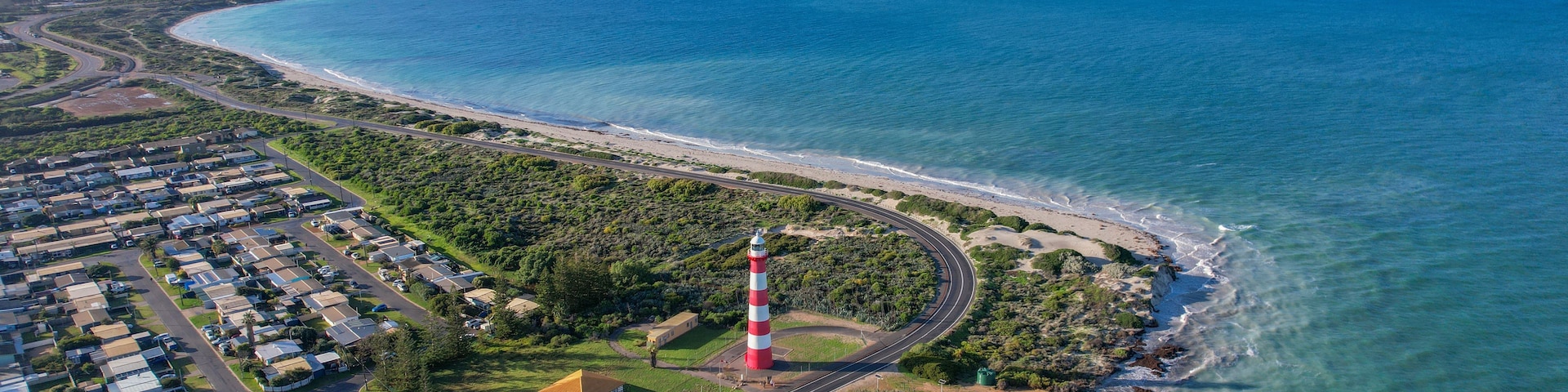 An aerial view of a coastal area with a lighthouse, a curved road, and a residential area with houses and greenery.