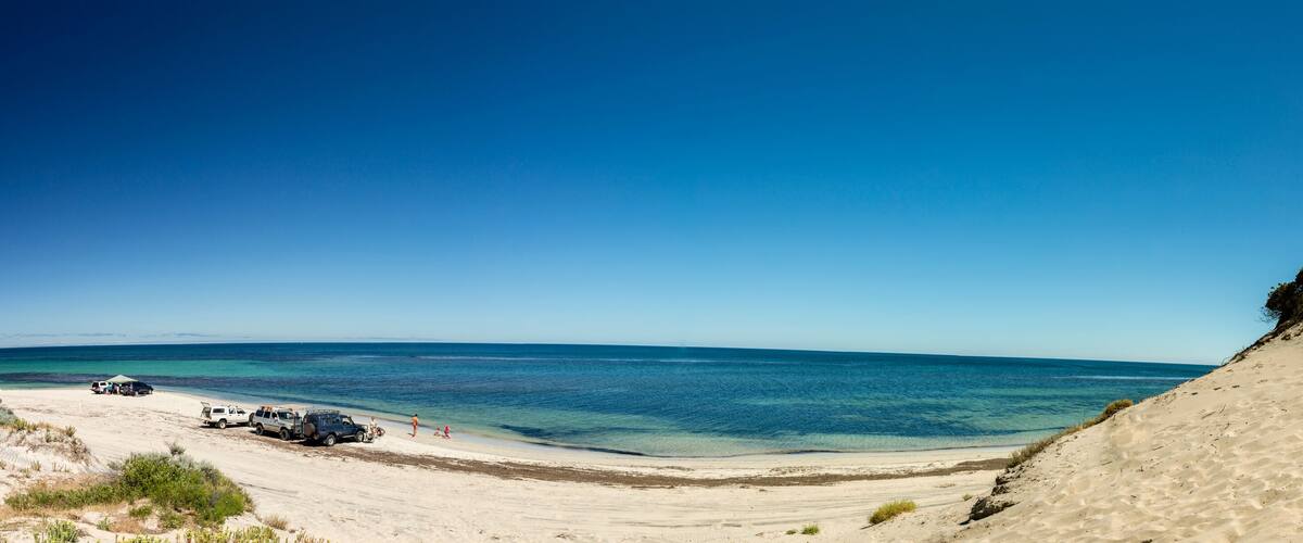 A coastal landscape with a clear blue sky, sandy beach, and a few vehicles parked near the shore.