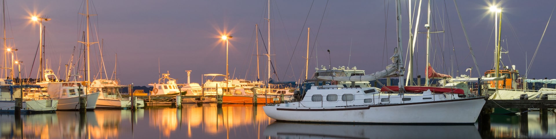 Un port de plaisance au crépuscule, où de nombreux voiliers sont amarrés, et leurs coques reflètent les lumières sur l’eau.