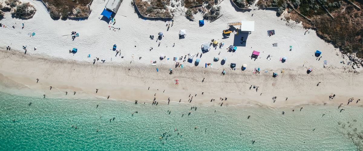 Vista aérea de una playa con aguas turquesas transparentes, gente tomando el sol y sombrillas de playa.