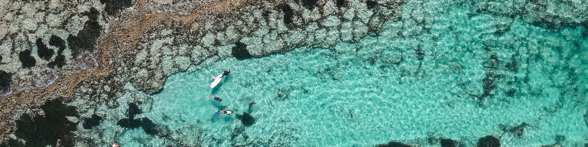 A coastal landscape with clear turquoise waters, rocky formations, and a person surfing.
