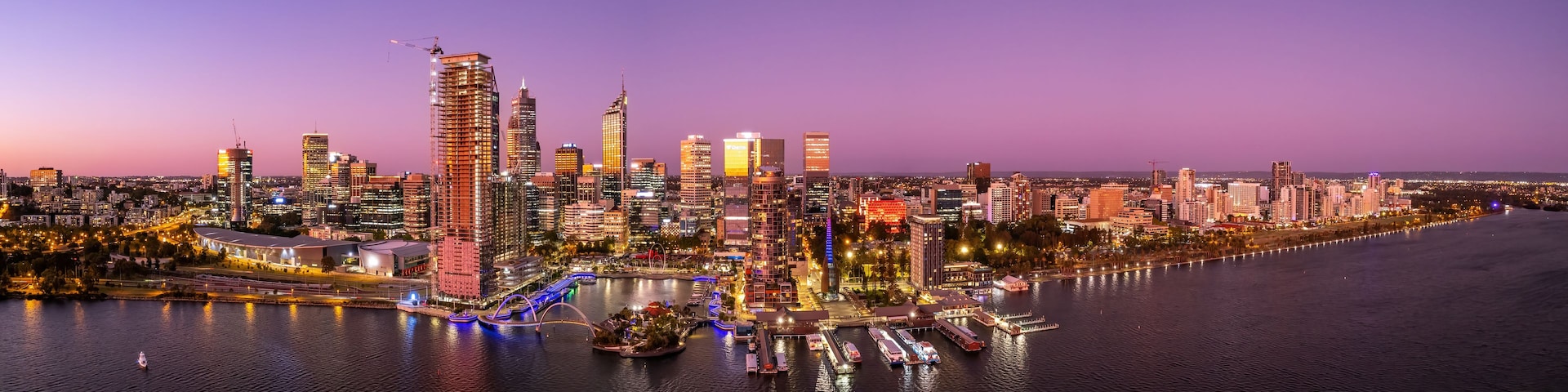 A city skyline at dusk with illuminated buildings and a river with boats.
