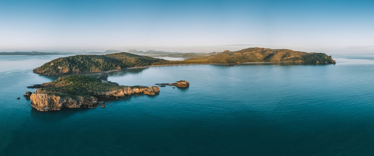A coastal landscape with a large island surrounded by calm blue waters, featuring rocky outcrops and dense greenery.