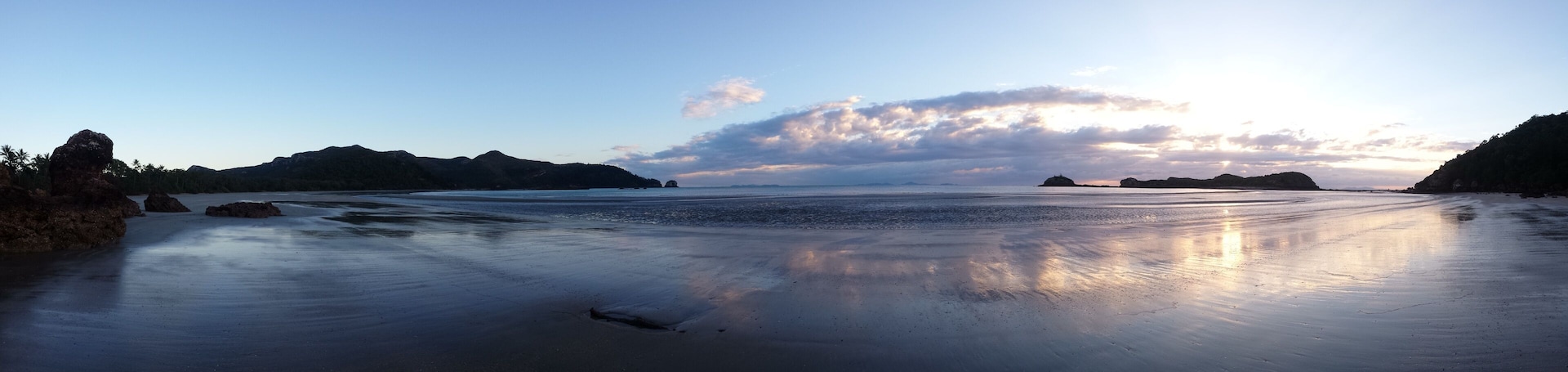 Uma praia com águas calmas, afloramentos rochosos e céu claro.