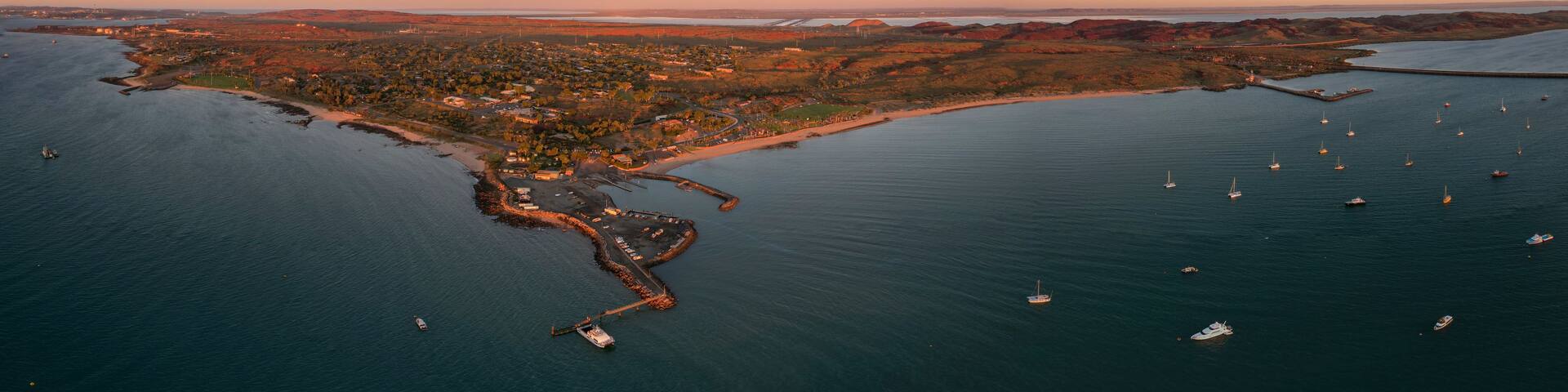 An aerial view of a coastal town with a marina, boats, and surrounding hills.