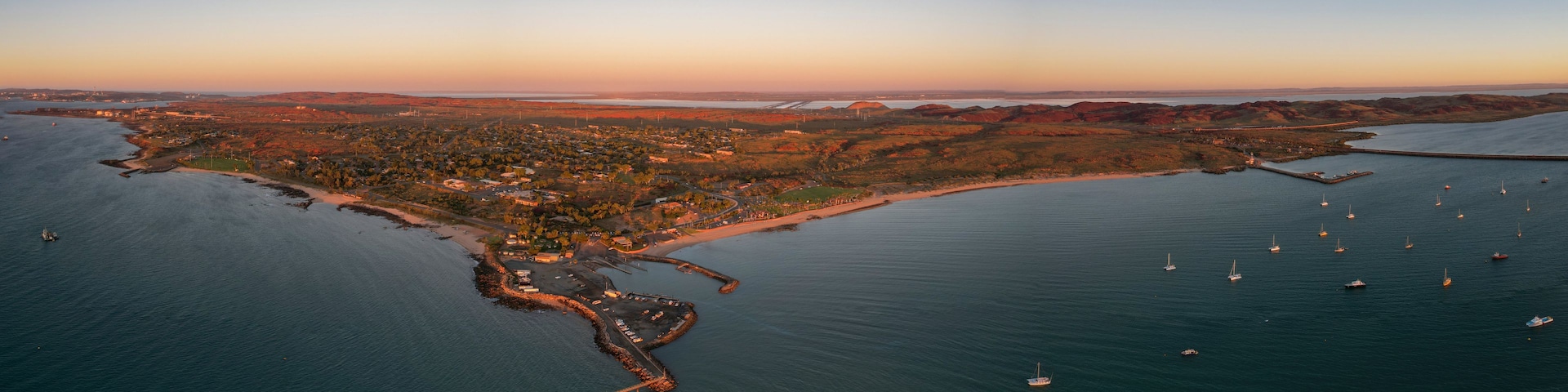 An aerial view of a coastal town with a marina, boats, and surrounding hills.