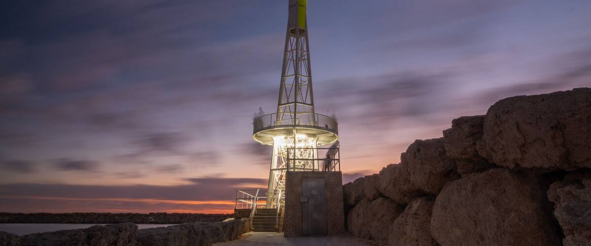 Farol ao entardecer, com céu claro e primeiro plano rochoso.