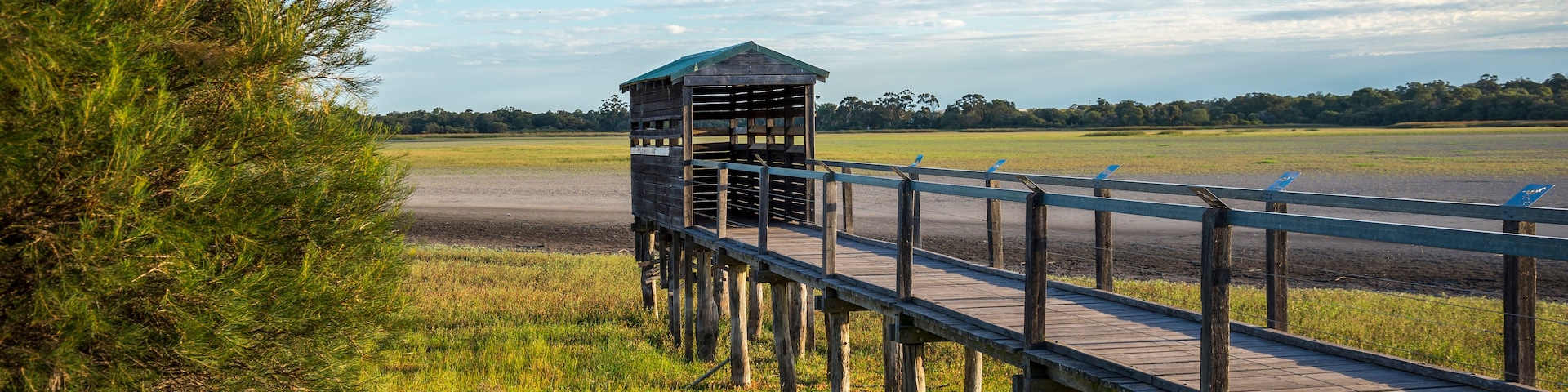 Un muelle de madera que conduce a un pequeño refugio con techo verde, rodeado de campos cubiertos de hierba y árboles bajo un cielo despejado.