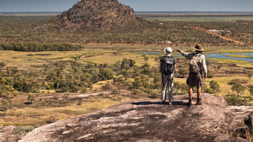 Two people standing on a rock overlooking a landscape with a hill and water bodies.
