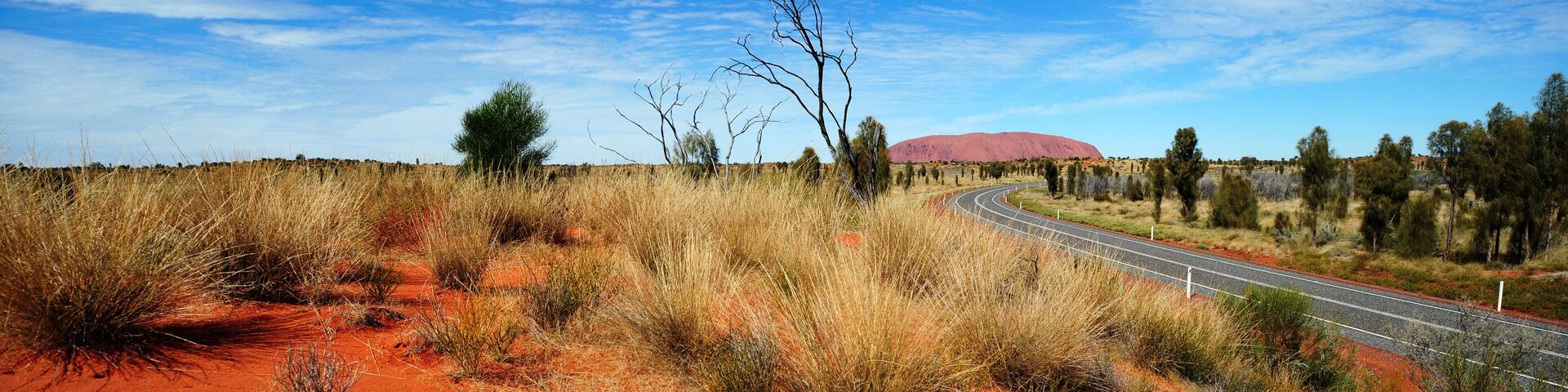 A winding road through a red desert landscape with sparse vegetation and a prominent red rock formation in the distance.