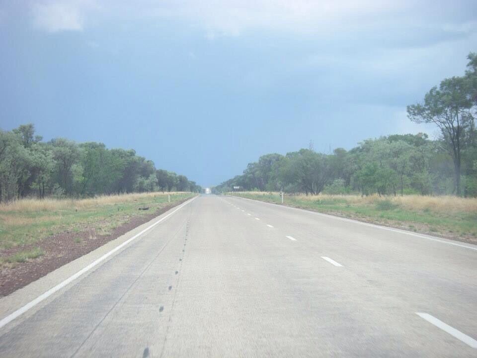 A straight highway flanked by trees and grasslands under a clear sky.