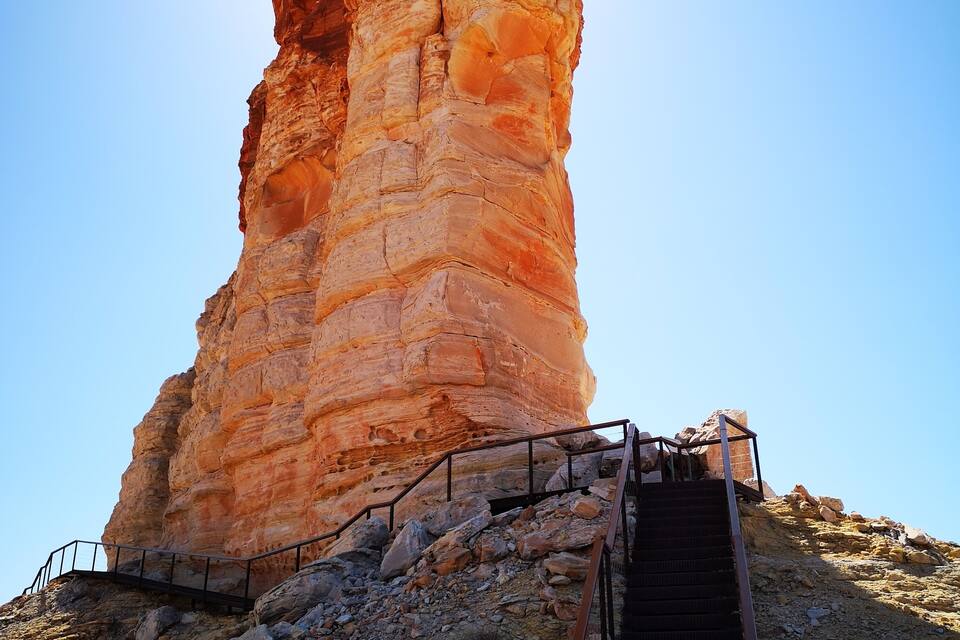 A tall, reddish-brown rock formation with a staircase leading up to it.