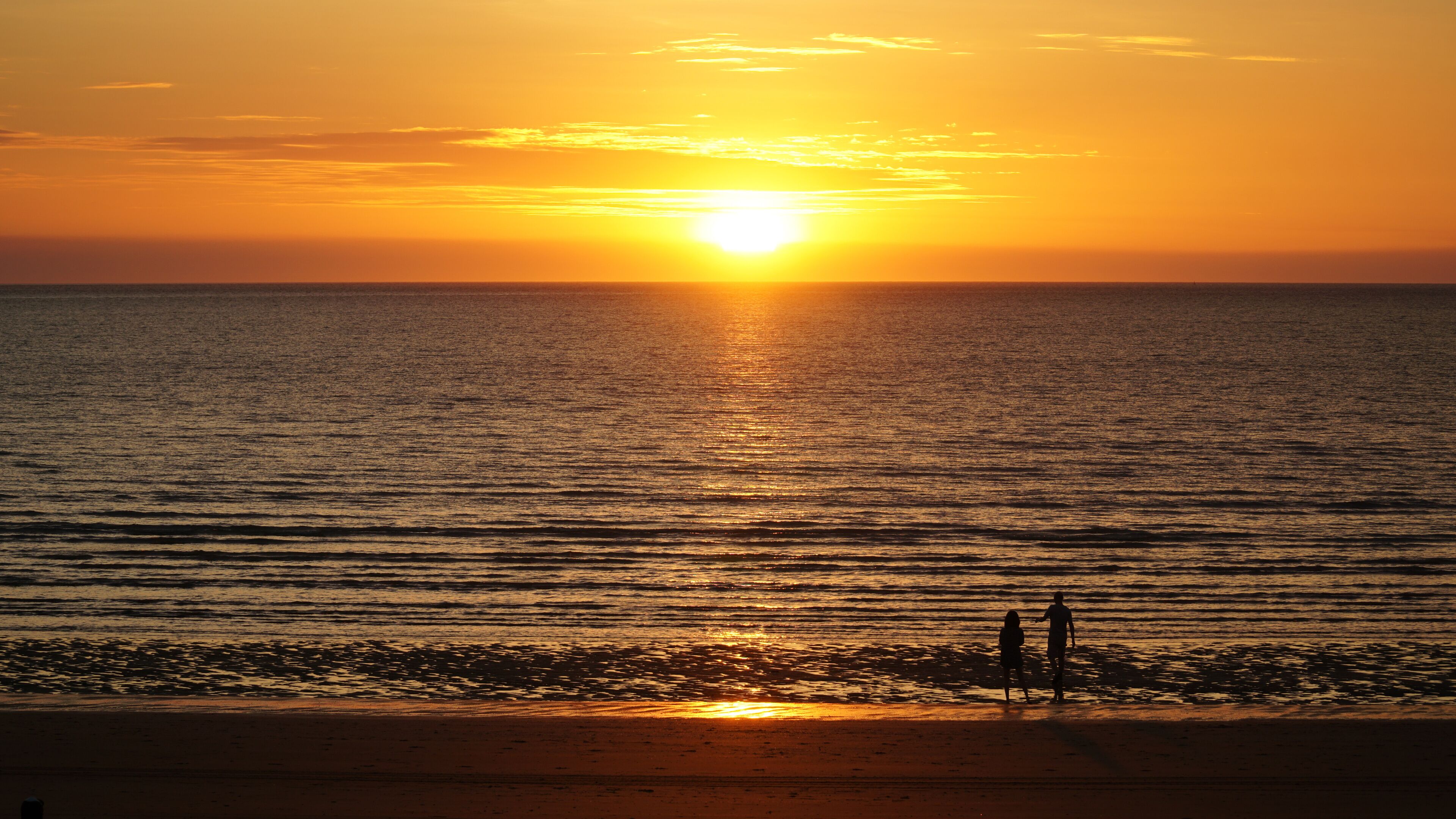 A sunset over a calm sea with a beach and two people walking.