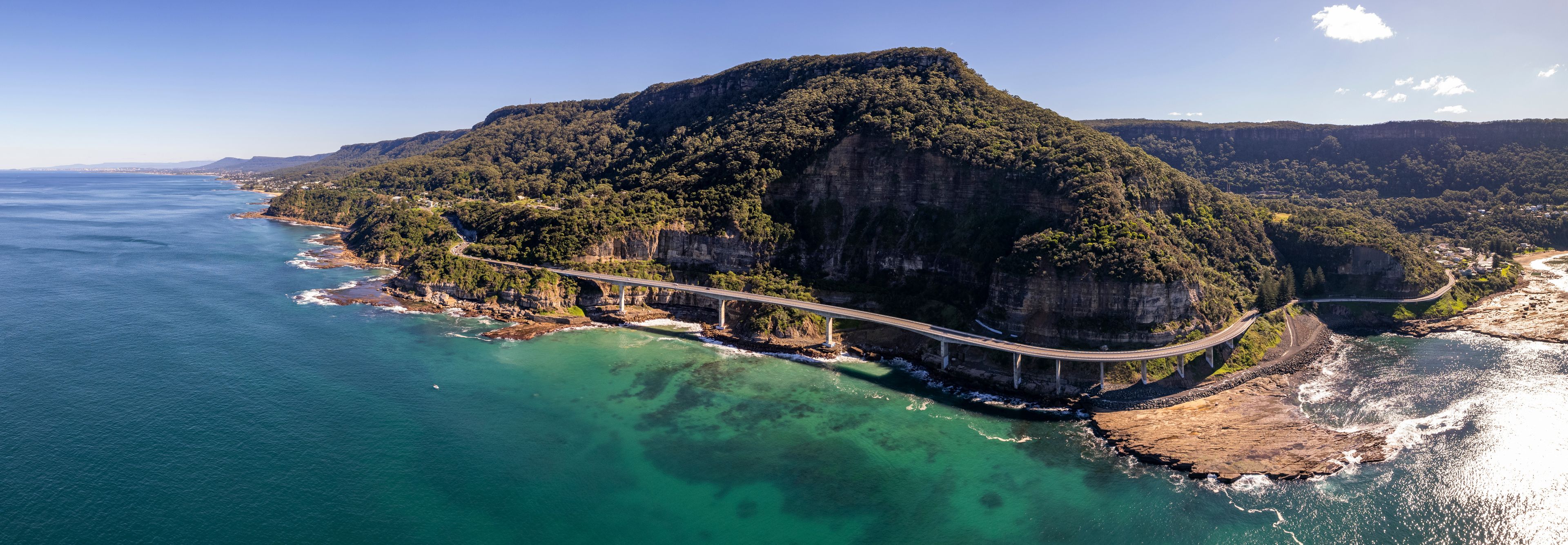 A coastal landscape with a prominent cliff, a bridge, and clear turquoise waters.