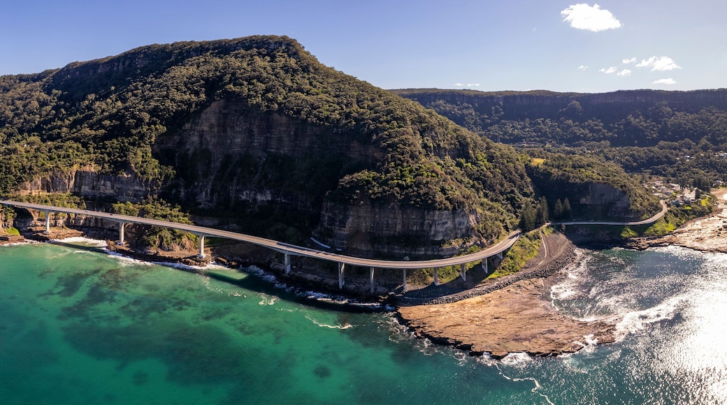A coastal landscape with a prominent cliff, a bridge, and clear turquoise waters.