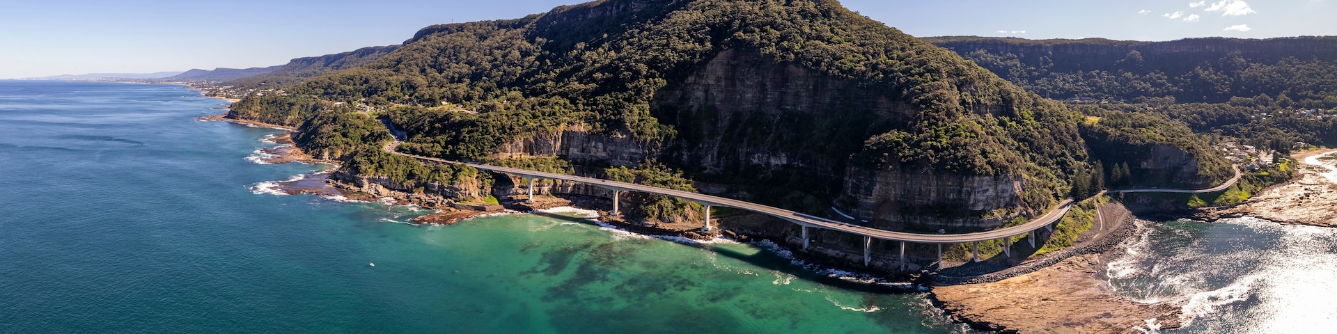 A coastal landscape with a prominent cliff, a bridge, and clear turquoise waters.