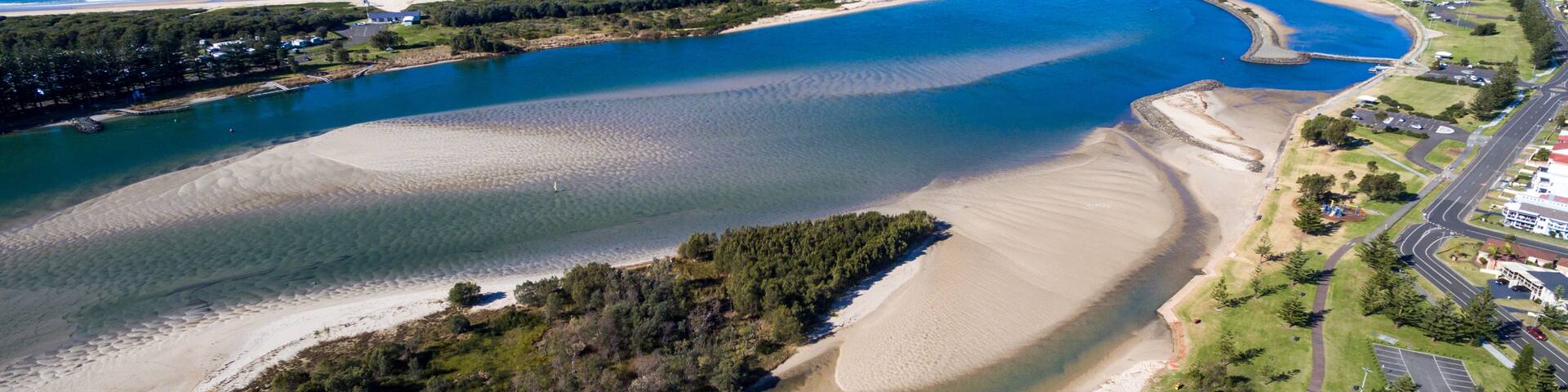 An aerial view of a coastal area with a large bay, sandy beaches, a small island with dense vegetation, and a road leading to a settlement.